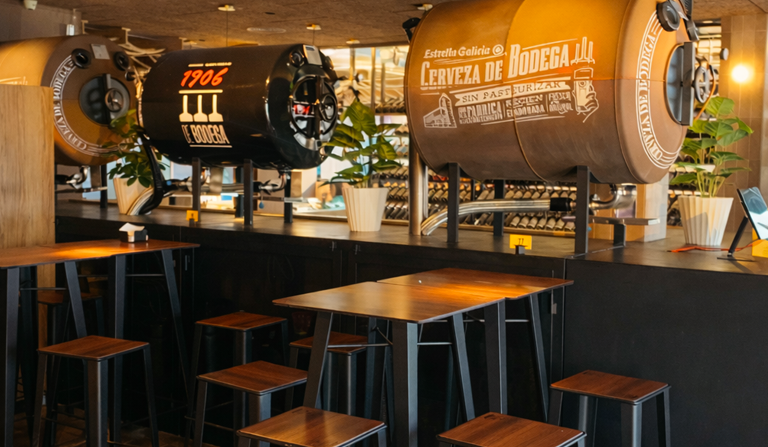 Zona interior d'OH TAPA! La Garriga amb taules altes, tamborets i dipòsits de cervesa de bodega en un ambient modern i acollidor.
